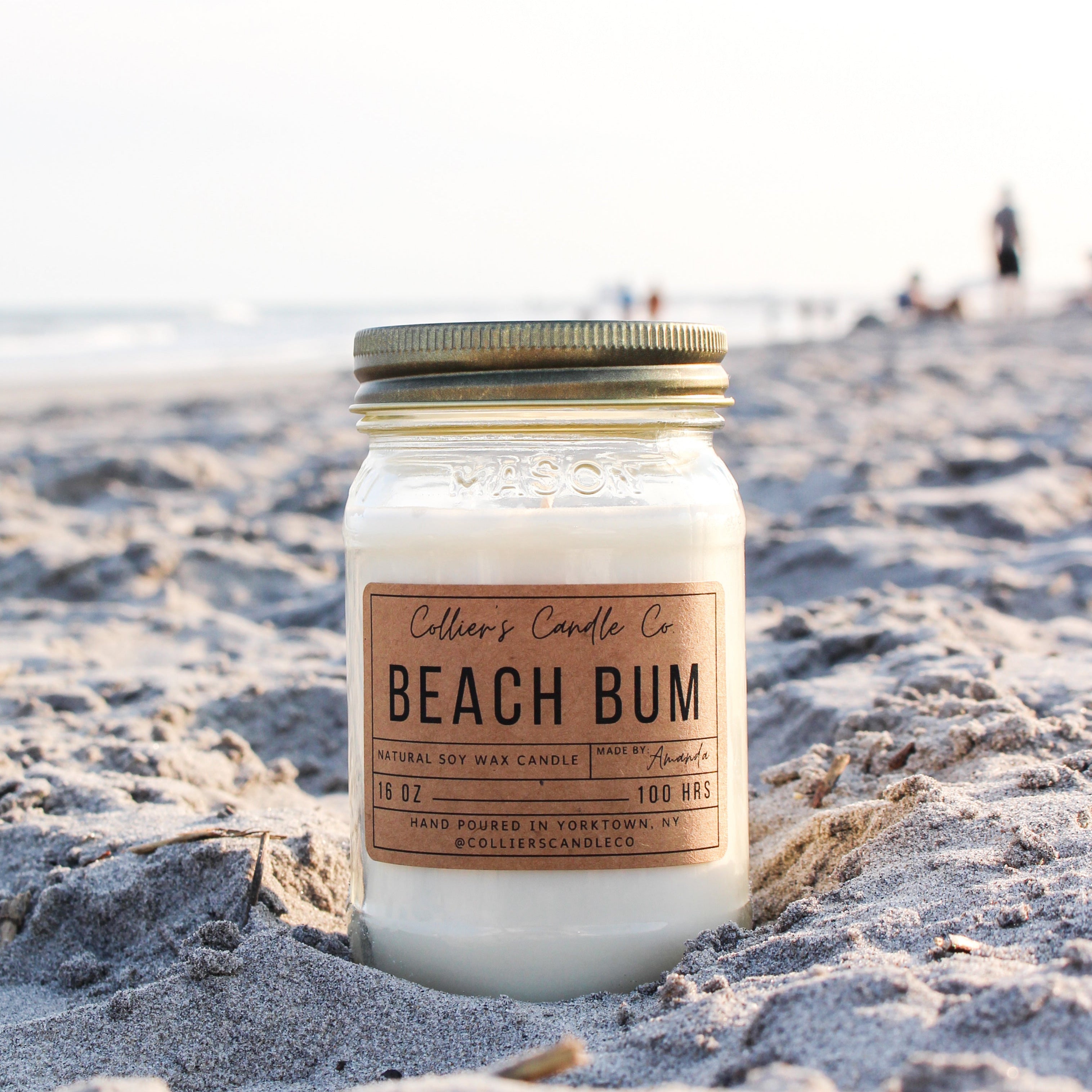 Candle labeled 'Beach Bum' on a sandy beach with ocean and people in the background