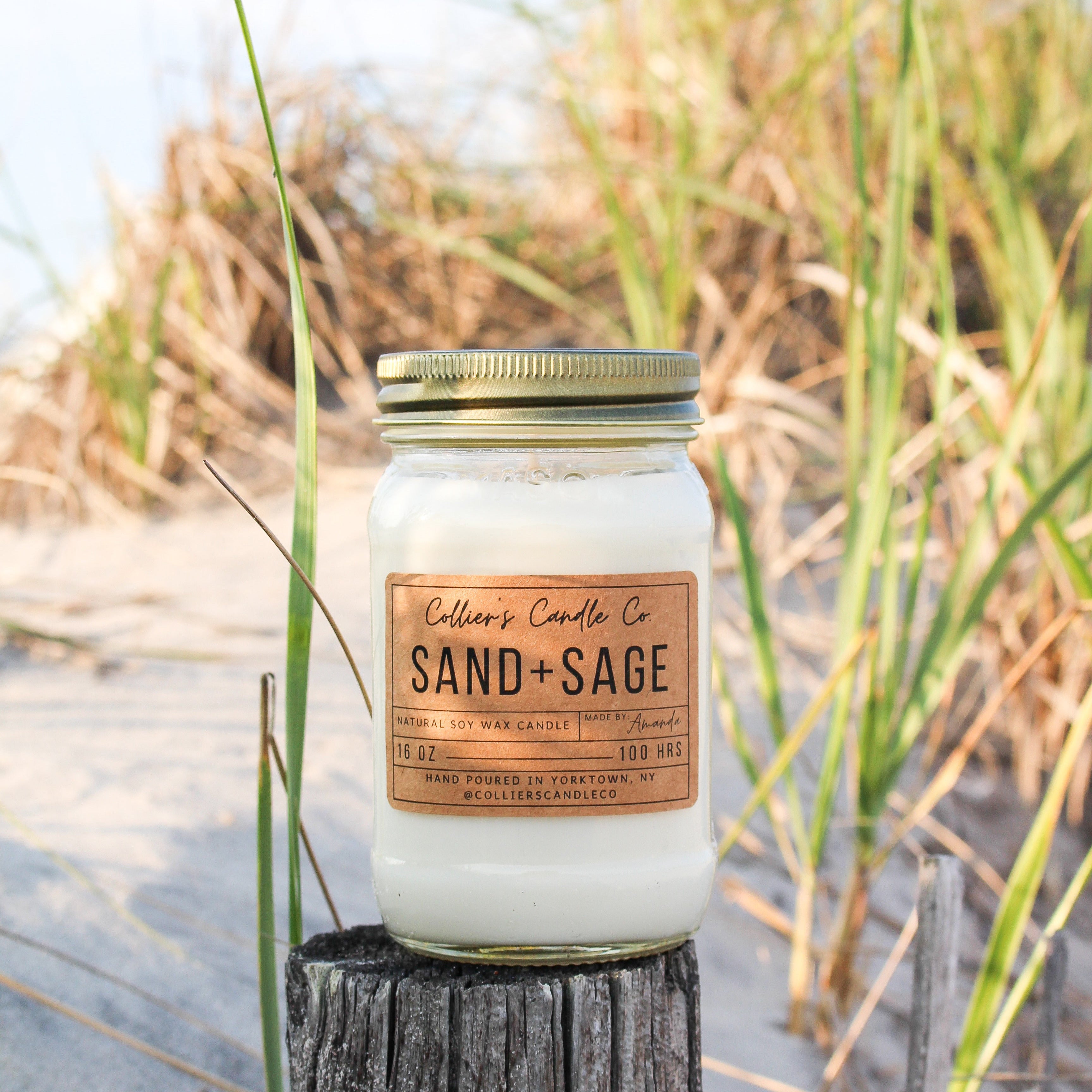Candle jar labeled 'Sand + Sage' on a wooden post with beach grass in the background