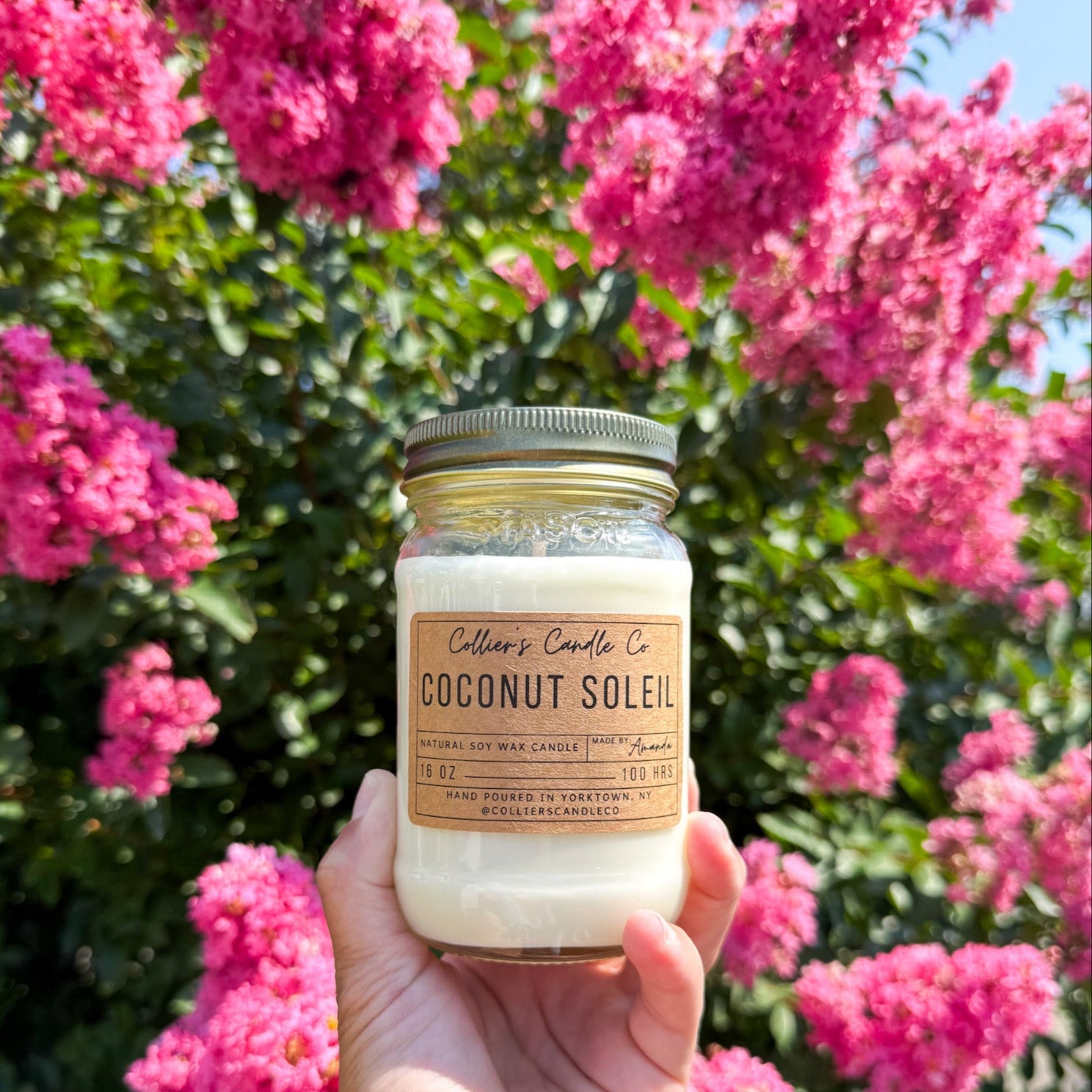 Hand holding a candle labeled 'Coconut Soleil' in front of pink flowers